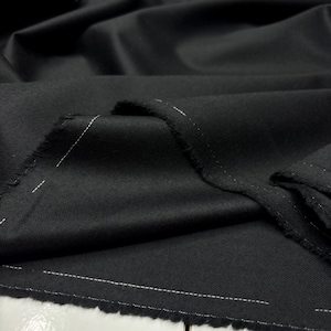 Op de afbeelding: Close-up van zwarte stof met witte stiksels. De stof is gevouwen en toont de textuur en de rafelige rand met de stiksels. Het materiaal lijkt een geweven textiel te zijn, mogelijk voor kleding of bekleding.