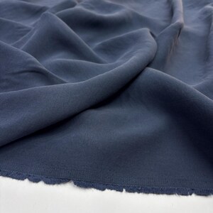 May include: A solid dark slate blue fabric with a matte finish, featuring soft folds and drapes that create natural shadows across the surface. The bottom edge of the textile shows a raw, frayed selvage with loose threads along the border.
