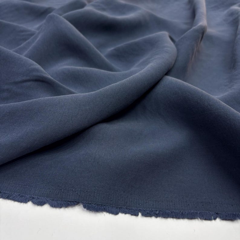 May include: A solid dark slate blue fabric with a matte finish, featuring soft folds and drapes that create natural shadows across the surface. The bottom edge of the textile shows a raw, frayed selvage with loose threads along the border.