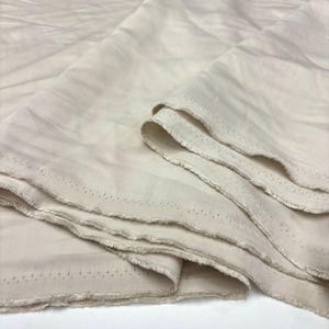 May include: A length of off-white fabric with a visible twill weave texture. The material is folded in multiple layers, showcasing the raw, unfinished edges with small, evenly spaced perforations along the selvage of the textile.