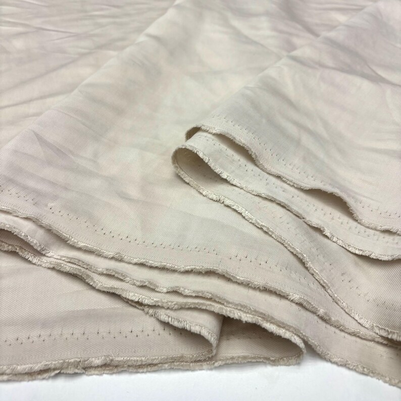 May include: A length of off-white fabric with a visible twill weave texture. The material is folded in multiple layers, showcasing the raw, unfinished edges with small, evenly spaced perforations along the selvage of the textile.