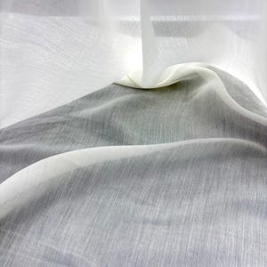 May include: Close-up of sheer white and grey fabric with a textured appearance. The fabric is draped, creating soft folds and shadows. The material appears lightweight and translucent, with a subtle sheen.
