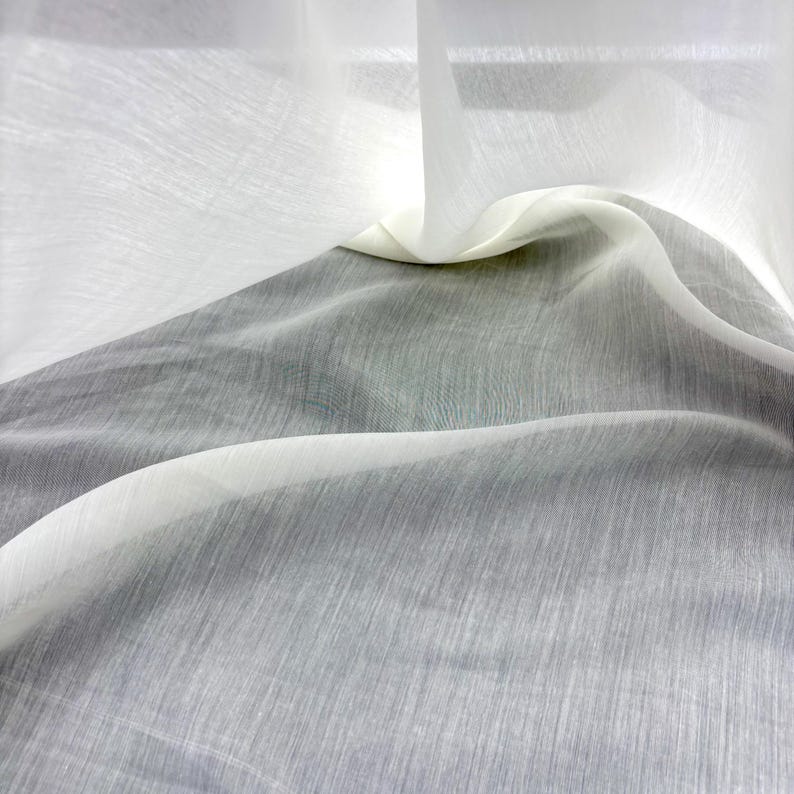 May include: Close-up of sheer white and grey fabric with a textured appearance. The fabric is draped, creating soft folds and shadows. The material appears lightweight and translucent, with a subtle sheen.