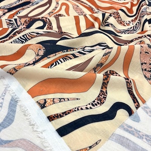 May include: Close-up of a fabric featuring a beige backdrop with a blend of animal print designs. The patterns incorporate zebra stripes, leopard spots, and snake scales in brown, black, and orange hues. The fabric is folded, revealing its texture.
