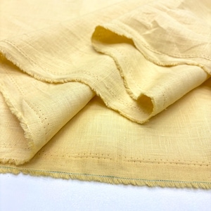 May include: Close-up of a folded piece of pale yellow fabric. The fabric has a textured appearance with visible weave and frayed edges. A row of small holes is visible near the edge, and a thin green line runs along the bottom edge.