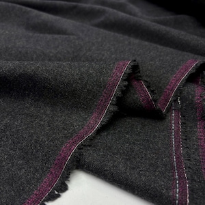 May include: Close-up of a dark gray fabric with a textured surface. The edges are frayed, revealing a burgundy stripe with white stitching. The fabric is folded, showing the texture and color variations.