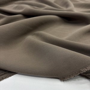 May include: A length of solid taupe-colored fabric featuring a distinct diagonal twill weave pattern. The material is draped in soft, flowing folds, showcasing a visible raw, frayed edge along the bottom corner of the textile piece.