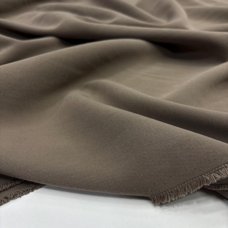 May include: A length of solid taupe-colored fabric featuring a distinct diagonal twill weave pattern. The material is draped in soft, flowing folds, showcasing a visible raw, frayed edge along the bottom corner of the textile piece.