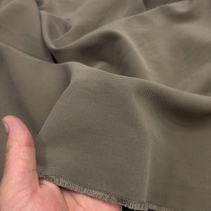 May include: A piece of solid taupe-colored fabric with a visible diagonal twill weave pattern. The material features a soft, matte finish and is shown with a frayed, unfinished edge, displaying the texture and drape of the textile material.