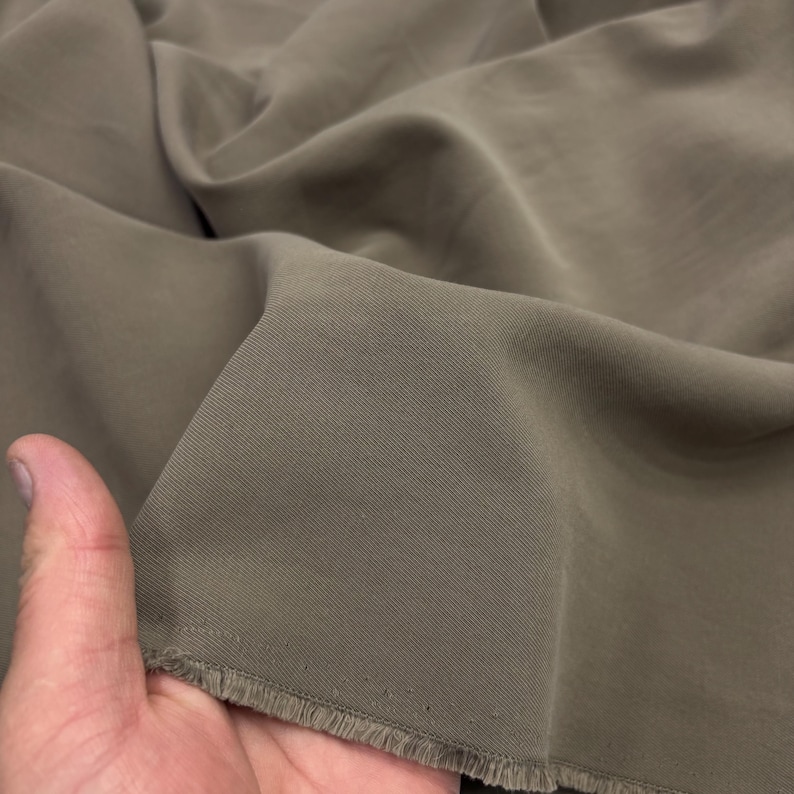 May include: A piece of solid taupe-colored fabric with a visible diagonal twill weave pattern. The material features a soft, matte finish and is shown with a frayed, unfinished edge, displaying the texture and drape of the textile material.