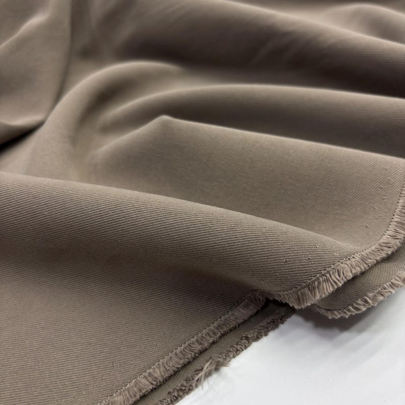 May include: A length of solid taupe-colored fabric featuring a distinct diagonal twill weave pattern. The material is draped in soft, flowing folds, showcasing a raw, unfinished edge with visible frayed threads along the bottom border.
