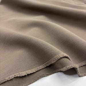May include: A length of solid taupe-colored fabric featuring a distinct diagonal twill weave pattern. The material is draped in soft folds, showcasing a textured surface and a raw, frayed edge along the bottom border of the textile piece.