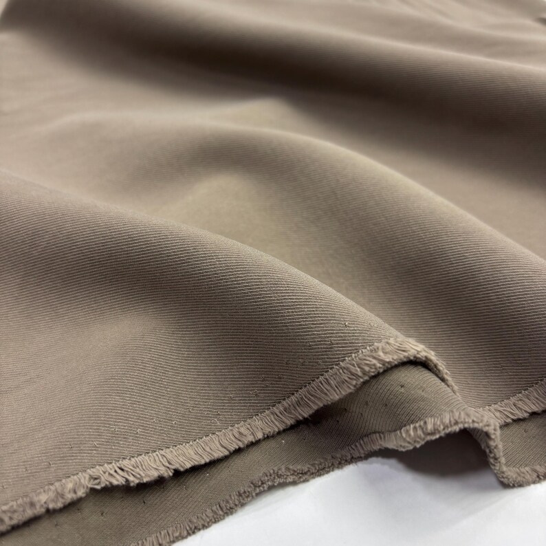 May include: A length of solid taupe-colored fabric featuring a distinct diagonal twill weave pattern. The material is draped in soft folds, showcasing a textured surface and a raw, frayed edge along the bottom border of the textile piece.