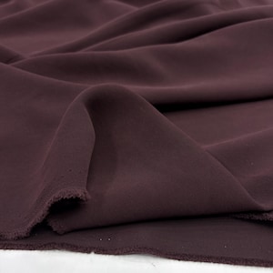 May include: A deep plum-colored fabric with a smooth, matte texture and a visible twill weave. The material is draped in soft, flowing folds, showcasing the consistent dark purple hue and the frayed, unfinished edges along the bottom border.