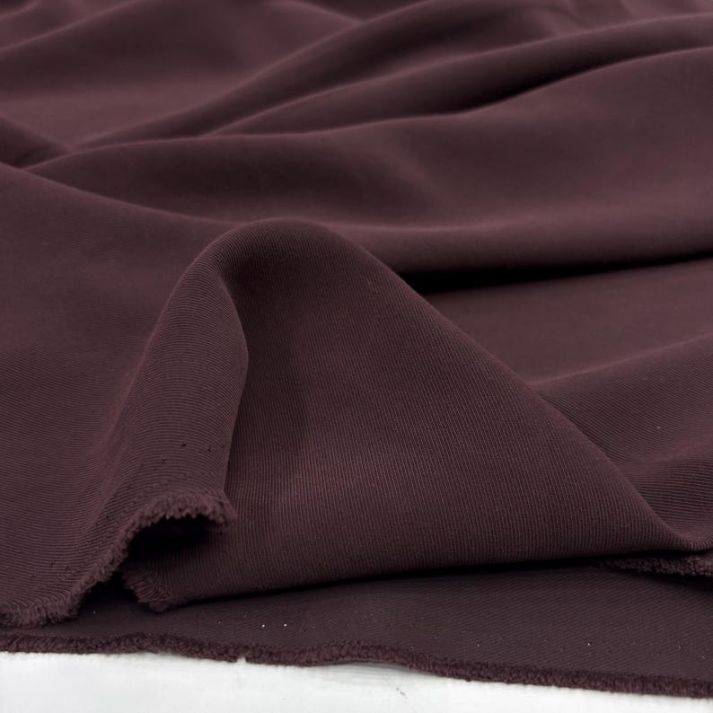 May include: A deep plum-colored fabric with a smooth, matte texture and a visible twill weave. The material is draped in soft, flowing folds, showcasing the consistent dark purple hue and the frayed, unfinished edges along the bottom border.