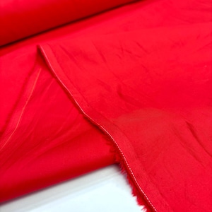 May include: A vibrant red fabric featuring a smooth, slightly reflective surface with subtle folds and creases. The material is shown in a folded layer, revealing a clean, straight finished edge with visible white stitching along the border.