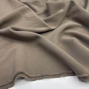 May include: A length of solid taupe-colored fabric featuring a visible twill weave texture. The material is draped in soft folds, creating deep shadows and highlights across the surface, with a raw, frayed edge visible along the bottom border.