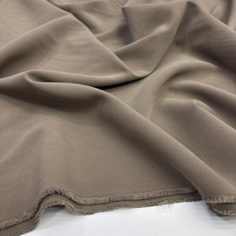 May include: A length of solid taupe-colored fabric featuring a visible twill weave texture. The material is draped in soft folds, creating deep shadows and highlights across the surface, with a raw, frayed edge visible along the bottom border.