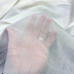 May include: Close-up of a sheer white fabric with a subtle, textured weave. The fabric is held up, allowing light to pass through, creating a translucent effect. The fabric appears soft and delicate, suitable for apparel or decorative projects.