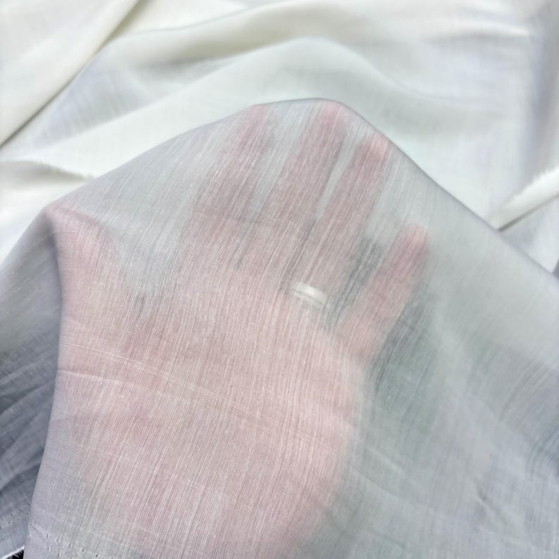 May include: Close-up of a sheer white fabric with a subtle, textured weave. The fabric is held up, allowing light to pass through, creating a translucent effect. The fabric appears soft and delicate, suitable for apparel or decorative projects.