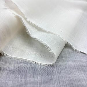 May include: Close-up of white fabric with a delicate, airy texture. The fabric is layered, showing a frayed edge and small, evenly spaced perforations. The material appears lightweight and slightly translucent, suggesting a soft, breathable textile.