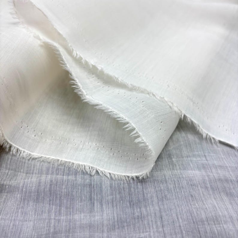 May include: Close-up of white fabric with a delicate, airy texture. The fabric is layered, showing a frayed edge and small, evenly spaced perforations. The material appears lightweight and slightly translucent, suggesting a soft, breathable textile.