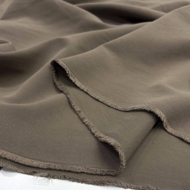 May include: A length of solid taupe-colored fabric featuring a visible twill weave pattern. The material is draped to show soft folds and ripples, with raw, unfinished edges displaying loose threads along the cut sections of the textile.