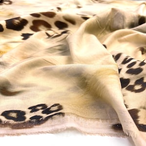 Puede incluir: Una tela ligera con un color base beige y un patrón de estampado de leopardo en marrón oscuro y negro. El material tiene una textura suave y drapeada con pliegues visibles y un borde crudo ligeramente deshilachado en el dobladillo inferior.