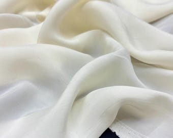 Puede incluir: Primer plano de una tela fluida de color blanco roto con un brillo suave y lustroso. La tela está drapeada en pliegues suaves, creando una sensación de movimiento y textura. Los bordes de la tela son visibles, mostrando un borde limpio y acabado.