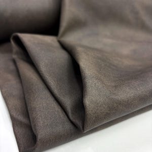 May include: A folded piece of dark brown fabric with a matte, textured surface. The material displays subtle variations in tone, creating a soft, mottled appearance across the draped folds of the textile, which is shown in a close-up view.
