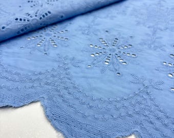 May include: Close-up of a light blue eyelet fabric. The fabric features intricate floral and geometric cut-out patterns. The edge of the fabric has a scalloped design. The fabric is folded over, showing the texture and detail of the material.