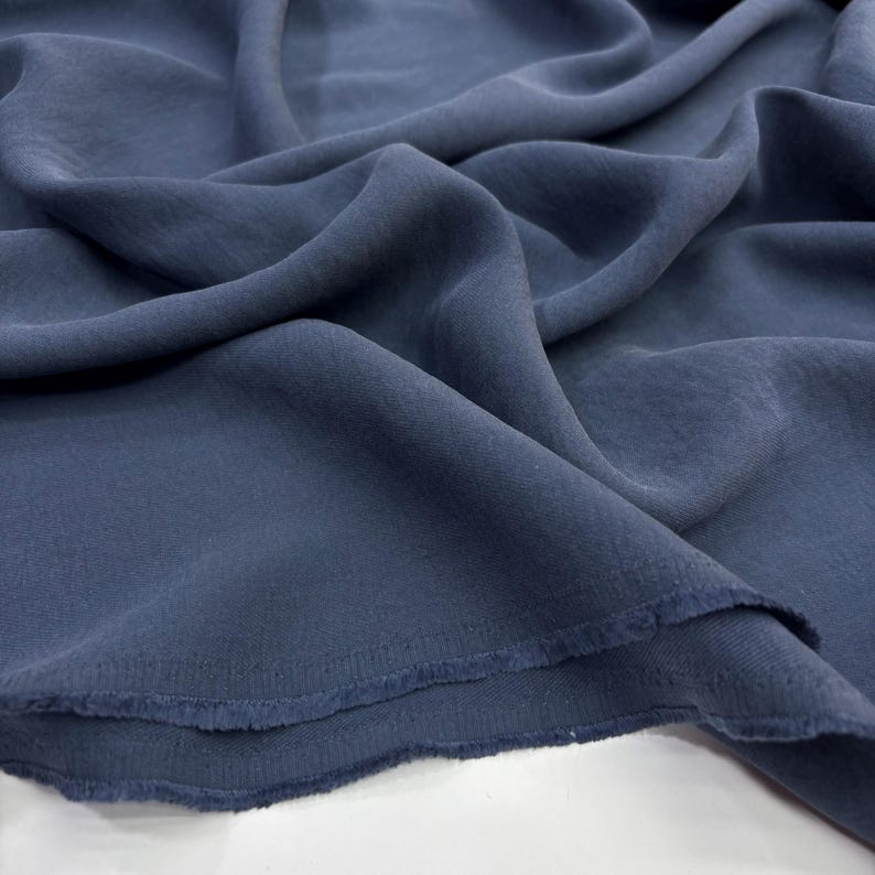 May include: A length of solid slate blue fabric with a soft, matte texture and a fluid drape. The material features a visible weave and raw, unfinished edges along the bottom, showcasing the textile's natural weight and flexible, folded composition.