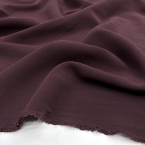 May include: A length of solid dark plum colored fabric with a smooth, matte texture. The material is draped in soft, flowing folds, creating deep shadows and highlights across the surface. The bottom edge shows a raw, frayed, and uneven hem.