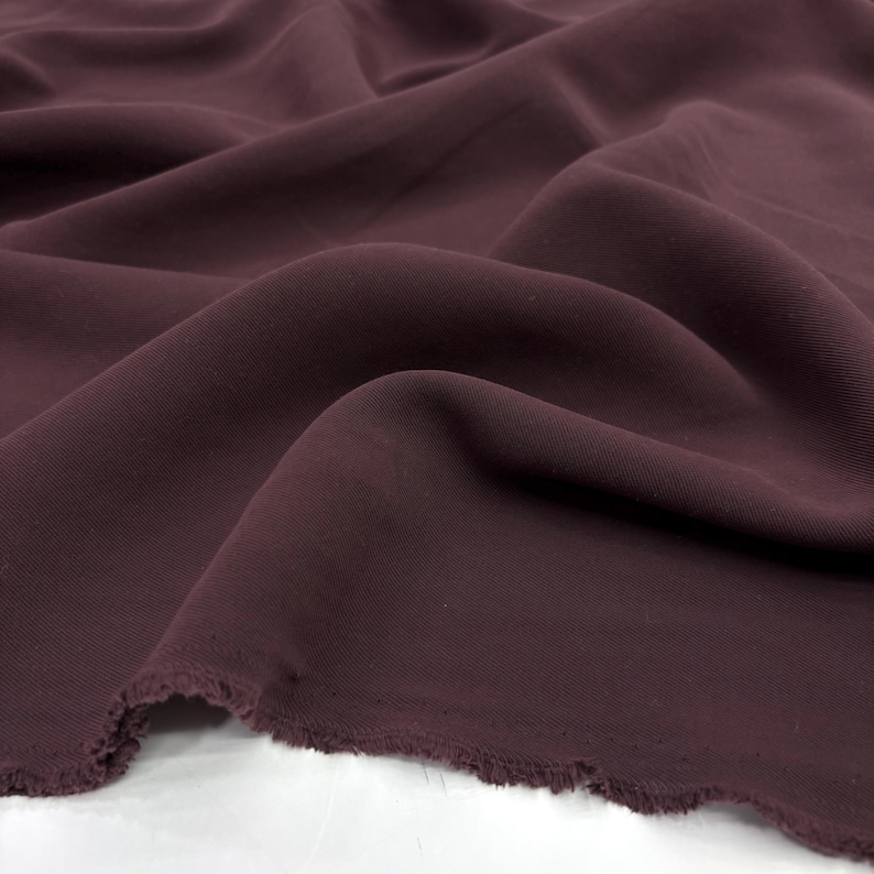 May include: A length of solid dark plum colored fabric with a smooth, matte texture. The material is draped in soft, flowing folds, creating deep shadows and highlights across the surface. The bottom edge shows a raw, frayed, and uneven hem.