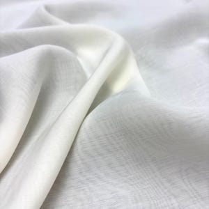 May include: A close-up shot of a flowing, off-white fabric with soft folds and subtle texture. The material appears lightweight and semi-transparent, with highlights and shadows creating depth. The fabric has a delicate, airy quality.