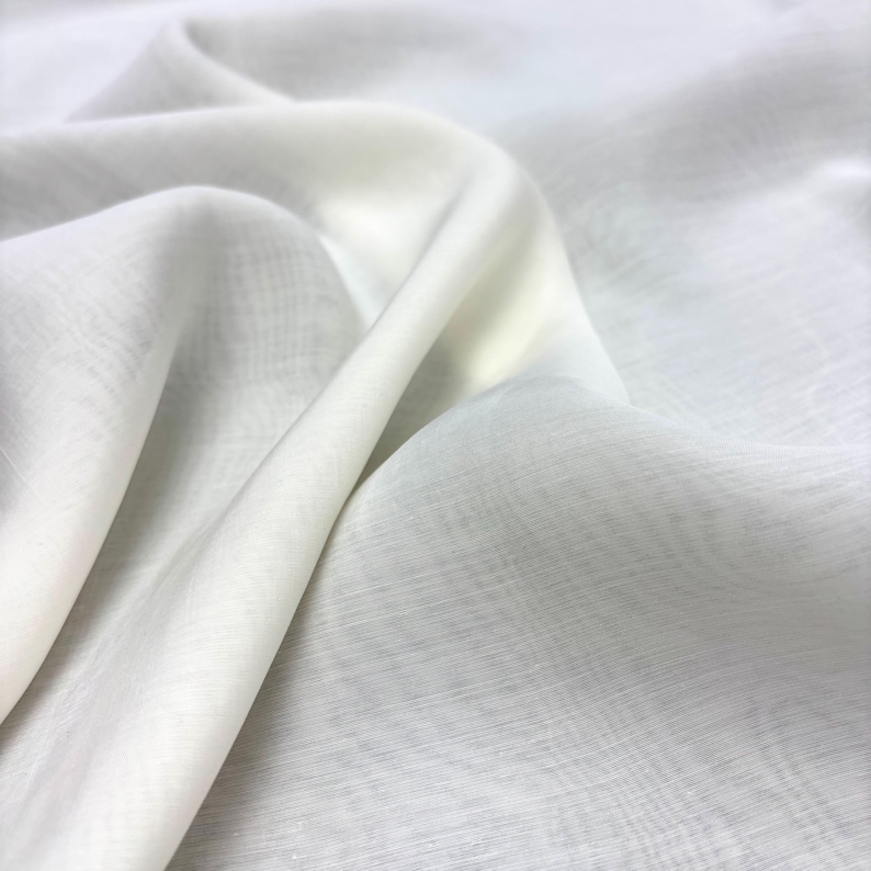 May include: A close-up shot of a flowing, off-white fabric with soft folds and subtle texture. The material appears lightweight and semi-transparent, with highlights and shadows creating depth. The fabric has a delicate, airy quality.