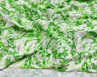 May include: White fabric with a vibrant green abstract pattern. The design features brushstroke-like markings in varying shades of green, creating a textured, artistic look. The fabric is draped, showing folds and shadows.