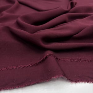 Könnte beinhalten: Ein Stück tiefburgunderroter Stoff mit einer glatten, matten Textur. Das Material ist in weichen, fließenden Falten drapiert, die natürliche Schatten und Lichter auf der Oberfläche erzeugen. Die Unterkante weist einen rohen, ausgefransten Saum auf.