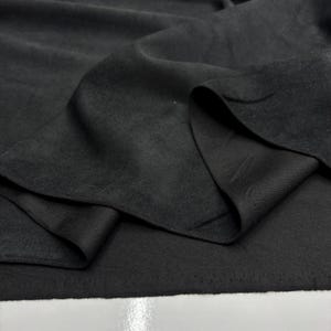 May include: Close-up of black fabric with a soft, matte texture. The fabric is draped, showing folds and layers. The material appears to be lightweight and flowing, with a subtle sheen. The image highlights the texture and depth of the dark textile.