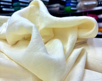 May include: Close-up of a folded, off-white fabric with a subtle texture. The fabric is draped, creating soft folds and shadows. The color is a creamy, pale yellow, suggesting a natural material like linen or cotton. The background shows other fabrics.