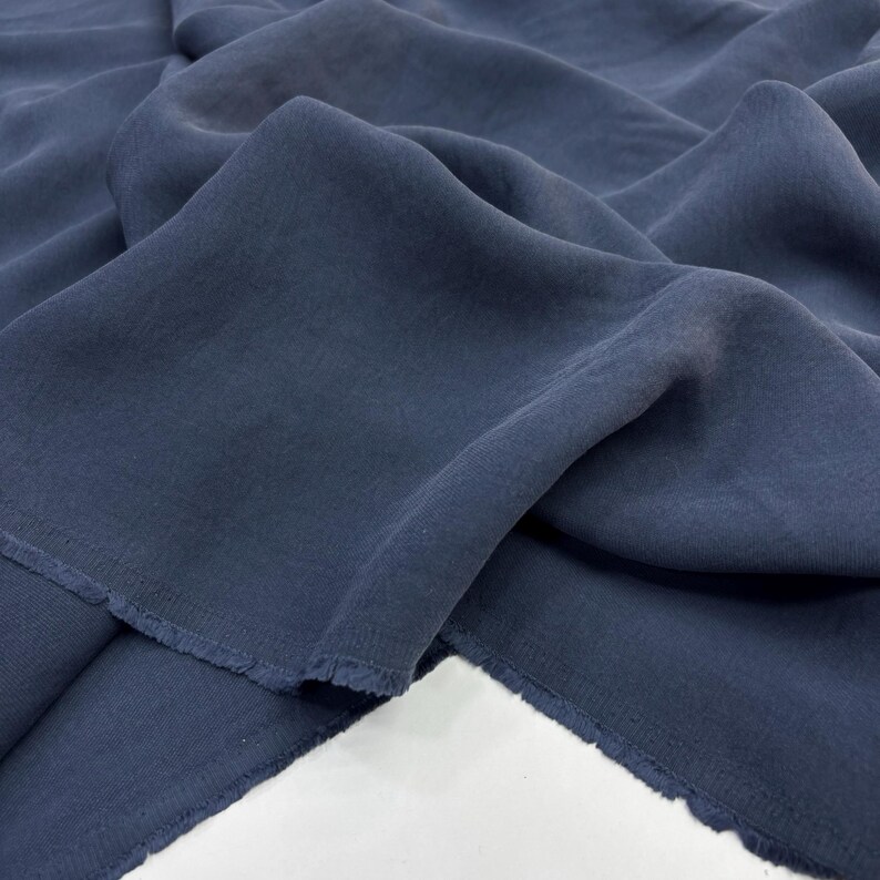 May include: A length of dark navy blue fabric with a soft, matte texture and visible woven grain. The material is draped in loose, flowing folds, showcasing the natural movement and raw, unfinished edges of the textile piece.