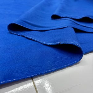 Puede incluir: Primer plano de una tela azul vibrante con un tejido texturizado. Los bordes est&aacute;n acabados con una costura blanca. La tela est&aacute; doblada, creando sombras suaves y resaltando la textura del material. El color azul brillante domina la imagen.