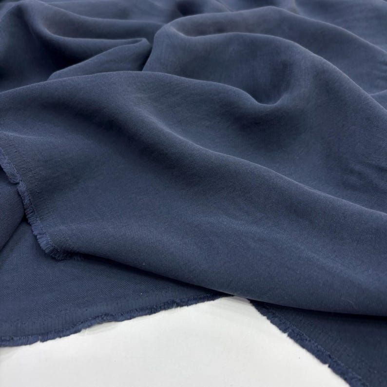 May include: A length of dark navy blue fabric with a soft, semi-sheer texture and a visible weave. The material is draped in loose, flowing folds, showcasing the matte finish and the raw, slightly frayed edges along the cut side of the textile.