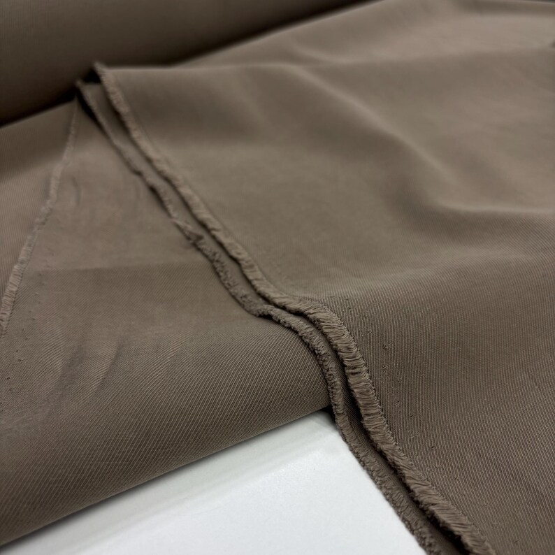 May include: A solid taupe-colored fabric featuring a distinct diagonal twill weave pattern. The material is folded to reveal a raw, frayed edge along the side, showcasing the texture and construction of the textile in a neutral, earthy brown tone.