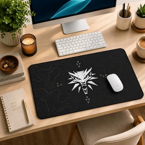 May include: A black rectangular mousepad with a white wolf head emblem and a topographic design. A white computer mouse sits on the pad. The desk also features a keyboard, monitor, and office supplies.