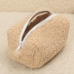 Può includere: Un beauty case rettangolare beige con un esterno soffice e strutturato. La borsa presenta una cerniera bianca e un interno in raso. La cerniera aperta rivela l'interno della borsa. La borsa è appoggiata su una superficie beige e strutturata.