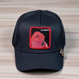 May include: Black baseball cap with a mesh back and a curved brim. The cap features a square patch with a flamingo illustration and the word "FLAMINGO" in white. A decorative button is on the side.