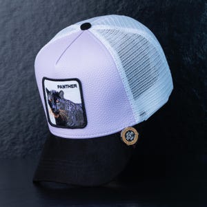 Può includere: Un cappellino da baseball con visiera nera e retro in rete bianca. Il pannello frontale è lilla chiaro con una toppa con la scritta "PANTHER" e un'illustrazione di pantera. Sul lato è presente una piccola spilla dorata.