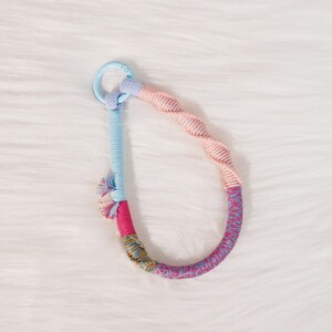 May include: A colorful keychain with a light blue clasp and a looped design. The loop is made of twisted pink and blue rope, with sections of pink, blue, and patterned fabric. The keychain has a tassel of pink, blue, and yellow threads.