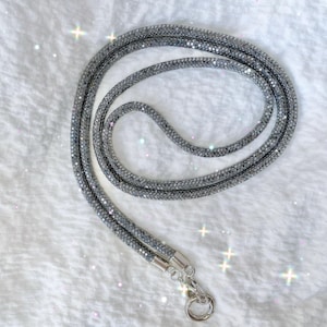 Puede incluir: Un cordón gris brillante con un cierre plateado. El cordón está cubierto de pequeños cristales reflectantes, creando un efecto brillante. El accesorio está diseñado para ser usado alrededor del cuello.